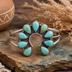 May include: A silver cuff bracelet featuring a horseshoe-shaped centrepiece adorned with nine teardrop-shaped turquoise stones. The metal band displays a twisted rope texture, and the turquoise stones exhibit natural brown matrix veining.