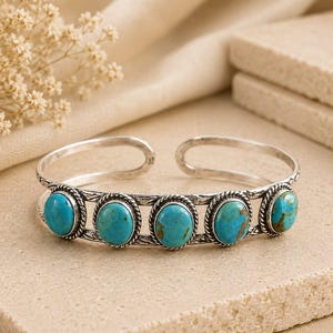 May include: A silver cuff bracelet featuring five oval-shaped turquoise stones set in twisted metal frames. The band has an open design with a textured, etched pattern along the metal surface, showcasing the bright blue and brown veined stones.