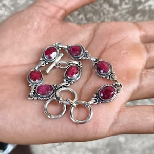May include: A silver bracelet featuring five oval-shaped, deep red faceted gemstones set in ornate, twisted metal frames. The piece includes a toggle clasp mechanism with a bar and two circular rings, showcasing a detailed, textured metallic finish.
