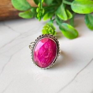 Può includere: Un anello d'argento con una grande gemma rosa brillante di forma ovale. La gemma è incorniciata da un bordo strutturato a forma di corda. L'anello è appoggiato su una superficie di marmo bianco, con foglie verdi sullo sfondo.