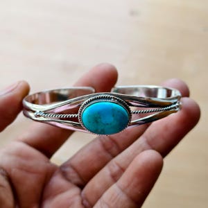Könnte beinhalten: Ein silbernes Armreif-Armband mit einem großen, ovalen Türkisstein. Der Türkis hat eine leuchtende blau-grüne Farbe und ist in einen detaillierten Silberrahmen gefasst. Das Armband hat ein schlichtes, elegantes Design.