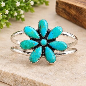 May include: A silver cuff bracelet featuring a central flower design composed of six teardrop-shaped turquoise stones and one small round turquoise stone in the centre. The metal band has a polished finish and a split design on both sides.