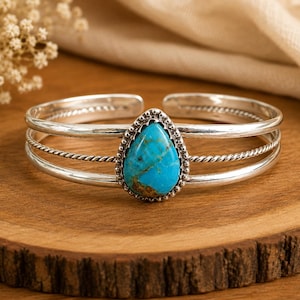 May include: A silver cuff bracelet featuring a central teardrop-shaped turquoise stone with brown matrix veining. The band consists of two smooth outer wires and a central twisted rope wire, with a beaded setting surrounding the turquoise stone.