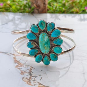 May include: A silver cuff bracelet featuring a large, central oval turquoise stone surrounded by twelve smaller, teardrop-shaped turquoise stones. The metal band is open at the back, and the stones display natural blue and green colour variations.