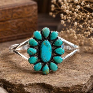 May include: A silver cuff bracelet featuring a large central oval turquoise stone surrounded by twelve smaller teardrop-shaped turquoise stones arranged in a floral pattern. The metal band is polished and split into two arms at the sides.
