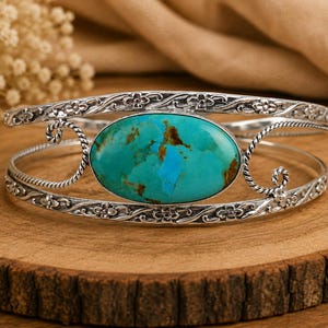 May include: A silver cuff bracelet featuring a large, oval-shaped turquoise stone with brown matrix patterns. The metal band is decorated with intricate floral engravings and twisted wire accents that frame the central gemstone setting.