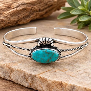 May include: A silver cuff bracelet featuring a central oval turquoise stone with brown matrix. The band consists of two smooth silver wires and two twisted silver ropes, accented by a small, fan-shaped silver ornament above the stone.