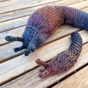 May include: Two hand-knitted slug figures crafted from variegated yarn in shades of charcoal gray, deep brown, and rust orange. The larger slug features two pairs of protruding tentacles, while the smaller slug has one pair of visible tentacles.