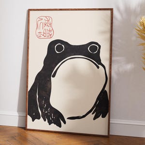 Peut inclure: Une affiche encadrée représentant une illustration minimaliste d'une grenouille noire sur fond crème. La grenouille a de grands yeux et une large bouche. Un motif rouge en forme de tampon est dans le coin supérieur gauche. L'œuvre est exposée sur un sol en bois.