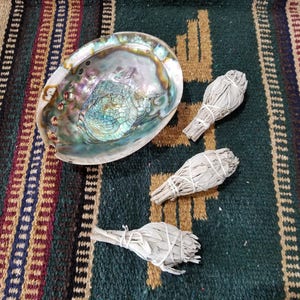 May include: Three white sage smudge sticks on a colorful striped rug with a large abalone shell.