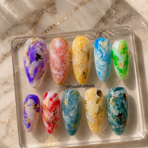 May include: A set of ten press-on nails arranged on a clear square display stand. Each nail features a unique marbled design in various colors including purple, pink, yellow, blue, green, and teal, accented with gold foil flakes and celestial motifs.