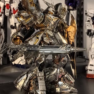 May include: A detailed, metallic silver and gold robot model with a large gun and shield. The robot has intricate armor plating and is posed in a dynamic stance. Other robot models are visible in the background, suggesting a collection of similar figures.