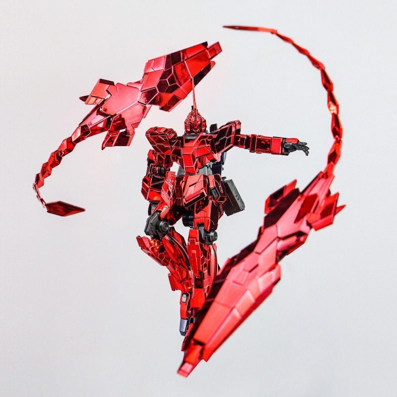 May include: A red robot action figure with articulated limbs and multiple weapon attachments. The figure is a metallic red color and has a detailed design. The robot is posed against a white background.