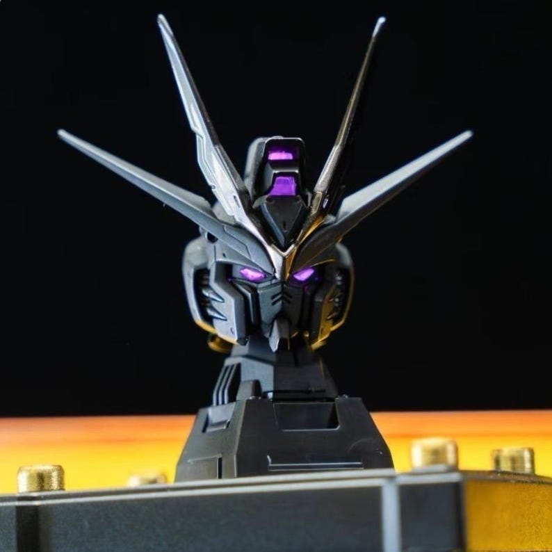 May include: A black and silver Gundam head model with purple accents. The model has sharp angles and a detailed design, with prominent antennae and glowing purple eyes. It is mounted on a black base, showcasing a futuristic aesthetic.