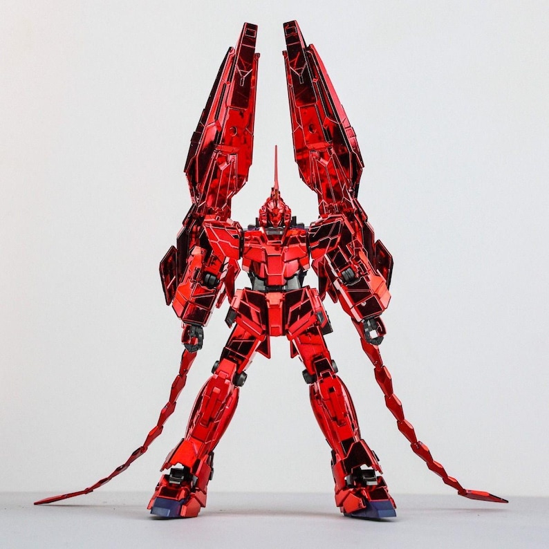 May include: A red robot action figure with large, angular wings and long, segmented tail-like appendages. The figure has a detailed design with a metallic finish, standing on a white surface. The robot's head features a prominent horn.