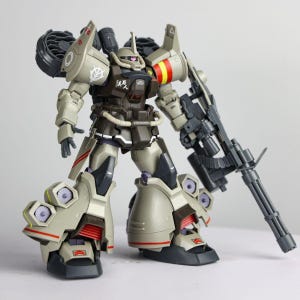 May include: A tan and dark brown robotic action figure featuring a large gatling gun, shoulder-mounted thrusters, and multiple leg-mounted vents. The figure displays white decals, red and yellow stripes on the shoulder, and a pink mono-eye sensor.