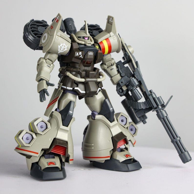 May include: A tan and dark brown robotic action figure featuring a large gatling gun, shoulder-mounted thrusters, and multiple leg-mounted vents. The figure displays white decals, red and yellow stripes on the shoulder, and a pink mono-eye sensor.