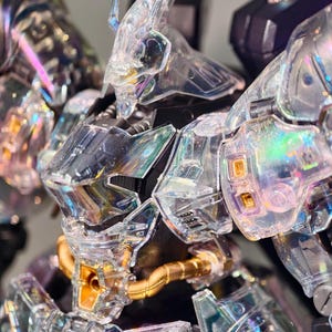May include: Close-up of a clear, iridescent robot model. The model features intricate details, including a transparent head, chest, and arms. Gold-colored tubes and accents are visible. The robot's design suggests a futuristic or science fiction theme.