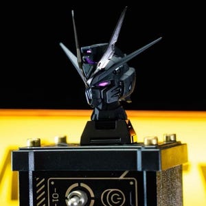 May include: A black and silver robot head model with purple accents, mounted on a black base. The model has sharp angles and details, with a futuristic design. The head is positioned above a black box with gold-colored knobs and a dial.