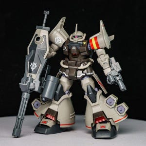 May include: A tan and dark brown plastic robot model kit featuring a large shield with a mounted gatling gun, a mono-eye sensor, and thruster details on the legs. The figure displays various decals, including a white emblem and Japanese characters.