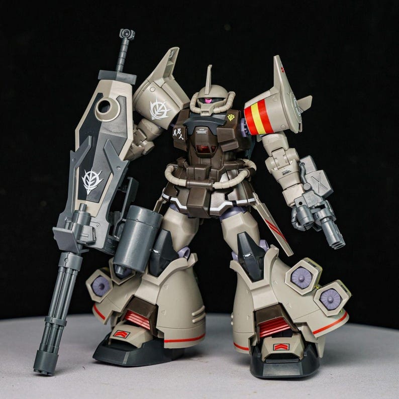 May include: A tan and dark brown plastic robot model kit featuring a large shield with a mounted gatling gun, a mono-eye sensor, and thruster details on the legs. The figure displays various decals, including a white emblem and Japanese characters.