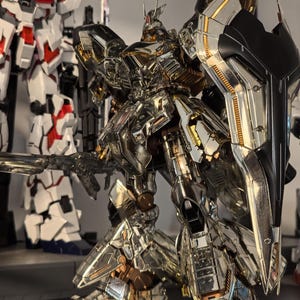 May include: A detailed, metallic silver and gold robot action figure with a large shield and weapon. The figure has intricate paneling and joint details, with a second robot figure visible in the background.