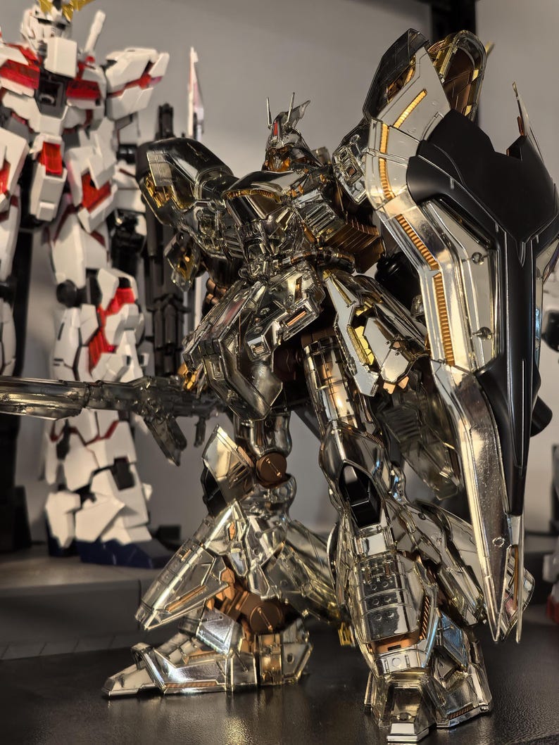 May include: A detailed, metallic silver and gold robot action figure with a large shield and weapon. The figure has intricate paneling and joint details, with a second robot figure visible in the background.