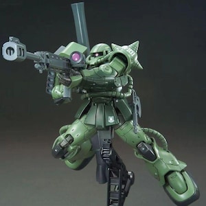Puede incluir: Un kit de modelo verde de un traje m&oacute;vil, posado en una postura de acci&oacute;n din&aacute;mica. La figura est&aacute; armada con un rifle grande y presenta un blindaje detallado. El modelo est&aacute; montado en un soporte de exhibici&oacute;n negro.