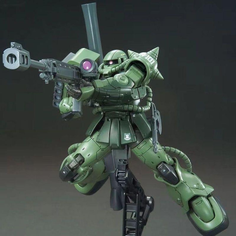 Puede incluir: Un kit de modelo verde de un traje m&oacute;vil, posado en una postura de acci&oacute;n din&aacute;mica. La figura est&aacute; armada con un rifle grande y presenta un blindaje detallado. El modelo est&aacute; montado en un soporte de exhibici&oacute;n negro.