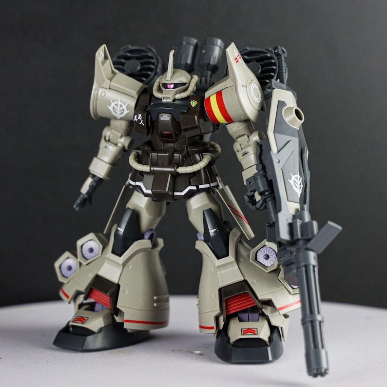 May include: A tan and dark brown robotic action figure featuring a mono-eye sensor, shoulder-mounted thrusters, and a large handheld gatling gun. The figure displays various decals, including red and yellow stripes on the shoulder armor.