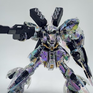 May include: A clear, iridescent model robot with a black gun. The robot has multiple black components and gold accents. The robot is posed with its legs spread and arms extended, as if in action. The model is set against a plain white background.