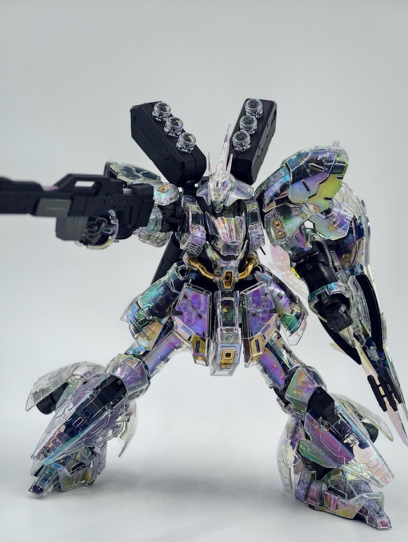 May include: A clear, iridescent model robot with a black gun. The robot has multiple black components and gold accents. The robot is posed with its legs spread and arms extended, as if in action. The model is set against a plain white background.