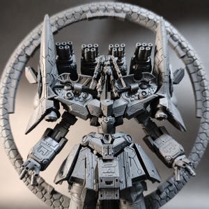 May include: A gray, detailed model of a futuristic robot with multiple cannons and a circular, segmented structure behind it. The robot has intricate armor plating and articulated arms, showcasing a complex design.