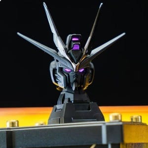May include: A detailed, black and silver Gundam model head with purple glowing eyes and accents. The model features sharp angles and a complex design, mounted on a black stand. The background is a gradient of black and yellow.