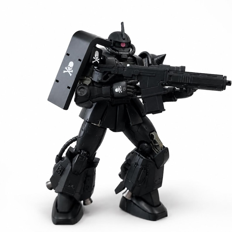 May include: A matte black articulated robot action figure featuring a large shoulder shield, a rifle, and a pink mono-eye sensor. The figure displays white skull and crossbones decals on the shield, shoulder, and hand, with metallic silver accents.