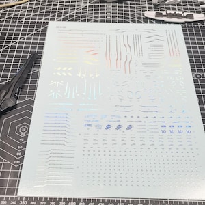 May include: A sheet of silver and iridescent decals for a model kit, featuring various lines, shapes, and symbols. The sheet is on a gray cutting mat with measurement markings and a partially assembled model piece.