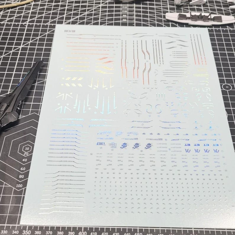 May include: A sheet of silver and iridescent decals for a model kit, featuring various lines, shapes, and symbols. The sheet is on a gray cutting mat with measurement markings and a partially assembled model piece.