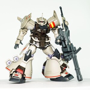 May include: A tan and dark brown robotic action figure featuring a mono-eye head, shoulder armor with red and yellow markings, and large leg thrusters. The figure holds a large dark gray multi-barreled weapon and displays white decals.