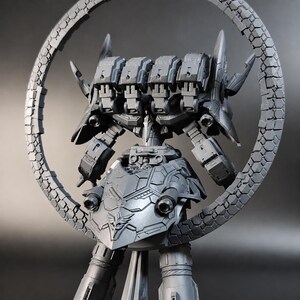 May include: A gray, detailed model of a futuristic robot with a circular frame. The robot has a complex design with multiple components, including a head with several rectangular sections and a shield-like chest. The circular frame is textured with a repeating pattern.