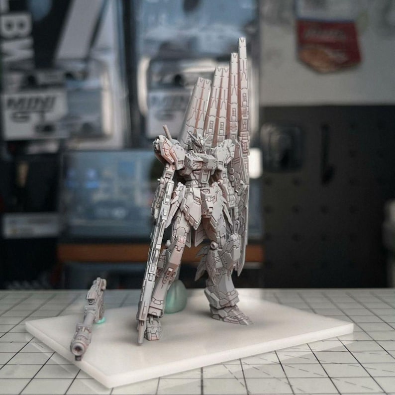 Pode incluir: Um modelo cinza detalhado de um rob&ocirc; Gundam, em p&eacute; sobre uma base branca. O modelo apresenta placas de armadura intrincadas, um escudo grande e um rifle. O rob&ocirc; est&aacute; posicionado com uma perna para a frente, sugerindo a&ccedil;&atilde;o. Uma arma menor &eacute; colocada ao lado.