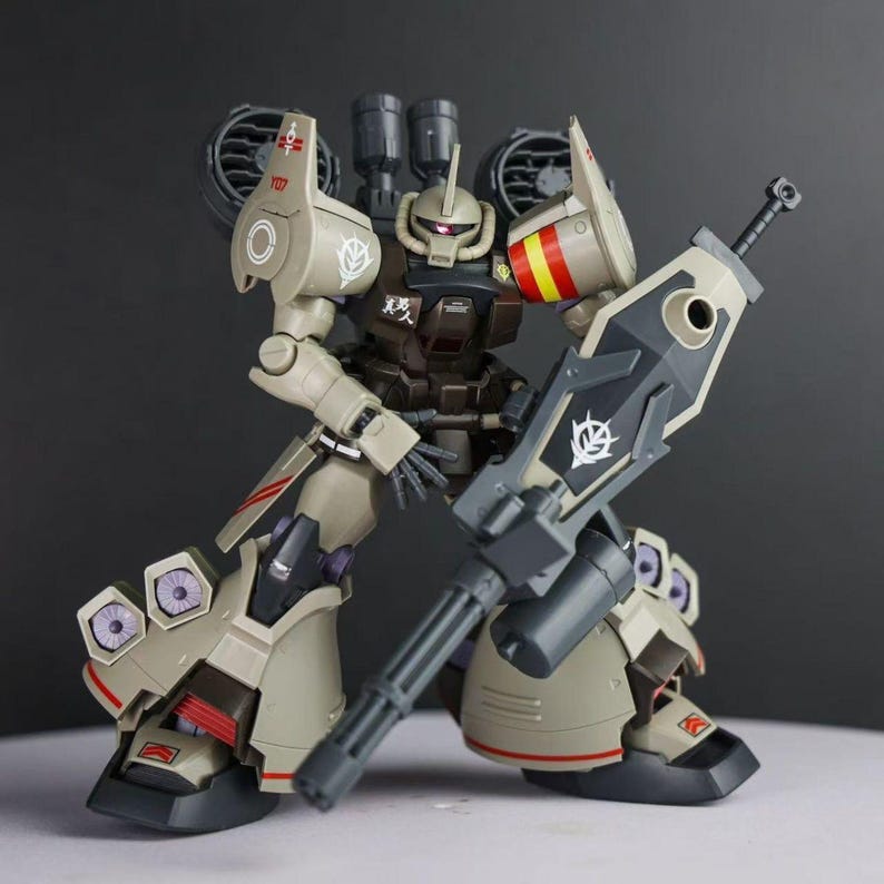 May include: A tan and dark brown robotic action figure featuring a large gatling gun, shoulder-mounted thrusters, and leg-mounted vents. The figure displays various decals, including a white emblem on the shoulder and red and yellow stripes.