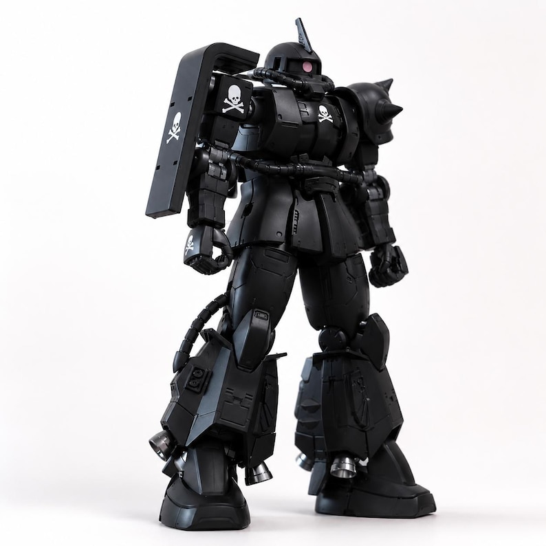 May include: A matte black Zaku robot action figure featuring a mono-eye sensor, a spiked left shoulder armor, and a large rectangular shield on the right arm. The figure is decorated with white skull and crossbones emblems on the shield, chest, and hand.