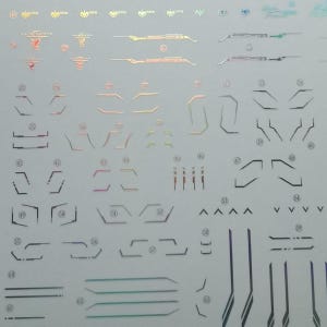 May include: A sheet of holographic stickers featuring various geometric shapes and lines in iridescent colors. The stickers include angular designs, arrows, and parallel lines, suitable for crafts or decoration.