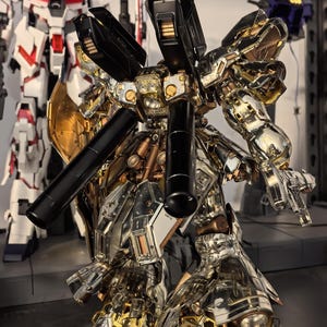 May include: A detailed model of a metallic gold, silver, and black robot with intricate mechanical details. The robot has large shoulder-mounted components and is posed with its arms extended. Another white and red robot is in the background.