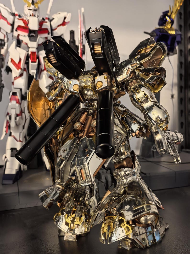 May include: A detailed model of a metallic gold, silver, and black robot with intricate mechanical details. The robot has large shoulder-mounted components and is posed with its arms extended. Another white and red robot is in the background.