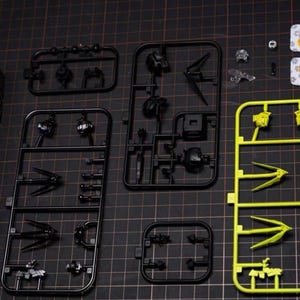 May include: A collection of unassembled model kit parts, including black and yellow plastic sprues. The parts are arranged on a black cutting mat, showcasing various components like mechanical figures and weapons. The image highlights the intricate details of the model kit.
