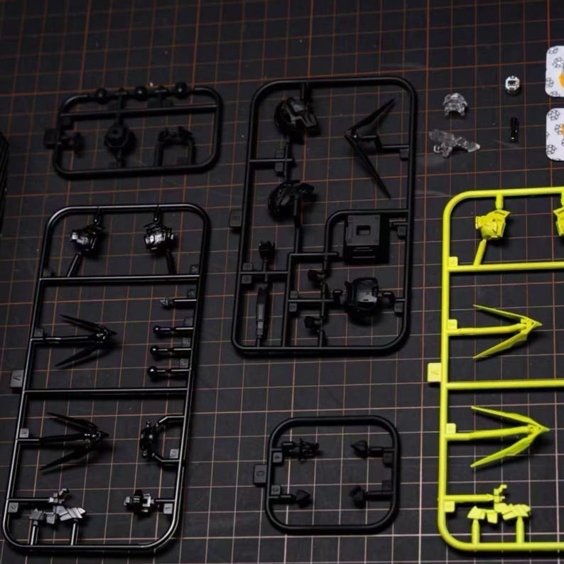 May include: A collection of unassembled model kit parts, including black and yellow plastic sprues. The parts are arranged on a black cutting mat, showcasing various components like mechanical figures and weapons. The image highlights the intricate details of the model kit.