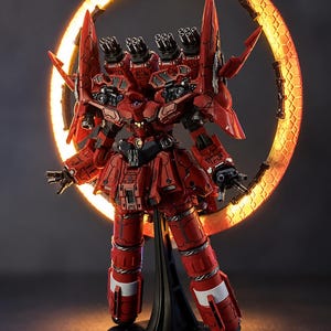 May include: A red and black robot model with multiple cannons and a glowing orange halo. The model is on a black stand, showcasing intricate details and a futuristic design. The robot has a skirt-like structure and white accents on its legs.