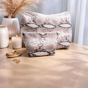 Può includere: Due trousse per cosmetici con stampa di serpente, una grande e una piccola, sono su una superficie di legno. Le borse hanno una chiusura con cerniera e una palette di colori neutri di bianco, grigio e marrone. Una candela accesa e oggetti decorativi sullo sfondo.