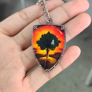 May include: A shield-shaped keychain featuring a vibrant sunset scene with a dark silhouette of a tree in the center. The keychain has a silver-toned metal frame and a matching silver chain link attachment at the top, showcasing a warm color palette.
